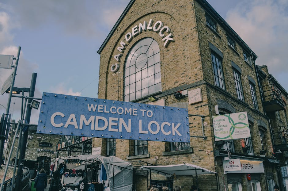 Exterior view of a three-story brick building with large arched windows and a prominent sign reading 'CAMDEN LOCK' at the top. In front of the building, a blue banner displays 'WELCOME TO CAMDEN LOCK'