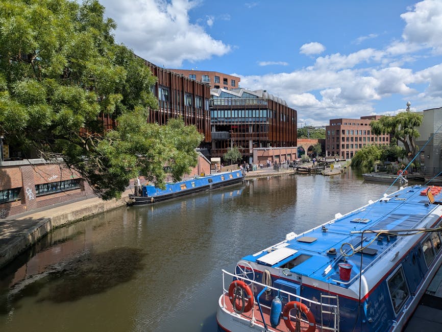 A wide view of a canal with several boats moored along the edge, including a prominent blue and white boat in the foreground equipped with safety rings, ropes, and a roof hatch. On the opposite side of the canal, there are modern multi-storey buildings with large windows and a mix of glass and brick facades, surrounded by mature trees with lush green foliage. The sky above is partly cloudy with patches of blue, and a paved walkway runs along the canal, where a few people can be seen walking or standing. This scene depicts urban river frontage typical of a city area, relevant to the context of Camden Town Removals' house and furniture transport services, with a focus on logistical considerations for home relocation and packing in an urban environment.