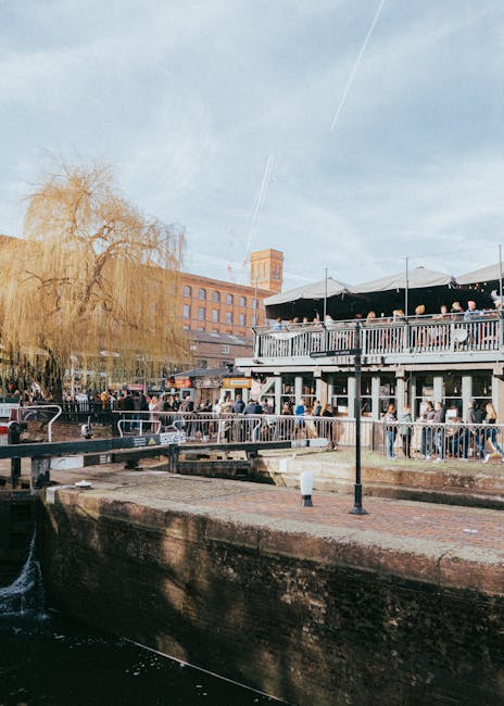 Image showing a busy outdoor scene along a canal or waterway in Camden, with a two-story building featuring large windows and an outdoor terrace filled with people seated at tables under umbrellas. A wooden railing borders the terrace, and a group of individuals appears to be engaged in activities related to house removals or moving preparations, such as carrying boxes or furniture. Several cardboard and plastic wrapping materials are visible, indicating packing or fragile item protection. A van is parked nearby on the pavement, with its rear open and partially visible, suggesting a loading or unloading process for a home relocation. A large tree with sparse foliage stands to the left, and the sky above is clear with a few contrails, contributing to the bright, daytime atmosphere. Camden Town Removals is associated with the scene, emphasizing logistics and transport involved in residential or commercial moves, particularly in the Camden area, consistent with the theme of house removals and furniture transport.